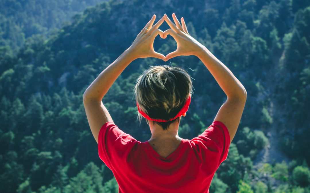 Young woman sitting towards mountains showing heart shape with her hands.