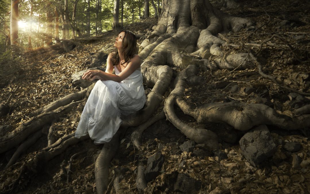 Lovely young lady wearing elegant white dress enjoying the beams of celestial light on her face sitting on the mighty roots of an ancient tree in enchanted woods